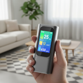 Compact handheld air quality monitor held in hand showing colorful readout screen, modern living room background